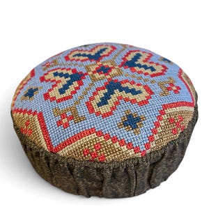 May include: A round pincushion featuring a cross-stitch pattern in light blue, dark blue, red, and tan on the top surface. The sides of the pincushion are made of a dark, textured, metallic-looking fabric with gathered pleats.