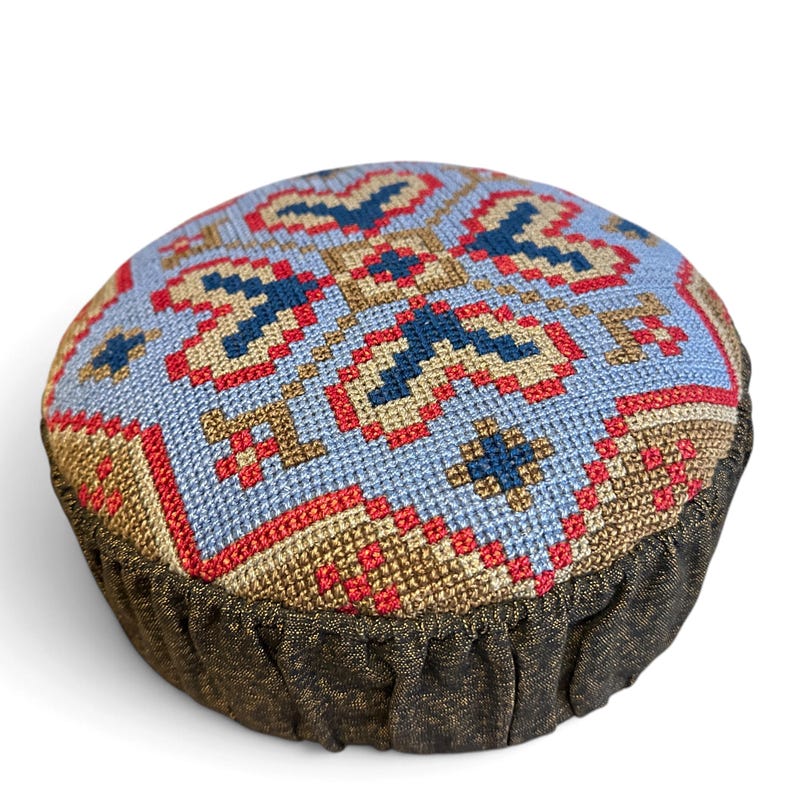 May include: A round pincushion featuring a cross-stitch pattern in light blue, dark blue, red, and tan on the top surface. The sides of the pincushion are made of a dark, textured, metallic-looking fabric with gathered pleats.