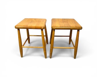 May include: Two wooden stools with square seats and four angled legs. The stools are made of light-colored wood and have a simple, classic design. The seats are approximately 12 inches wide.