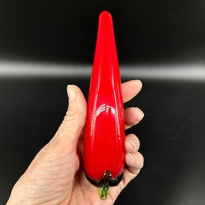 May include: A smooth, elongated red glass sculpture shaped like a chili pepper. The object features a glossy, vibrant red body that tapers to a point at the top, with a small, dark green glass stem detail at the base, reflecting light on its surface.