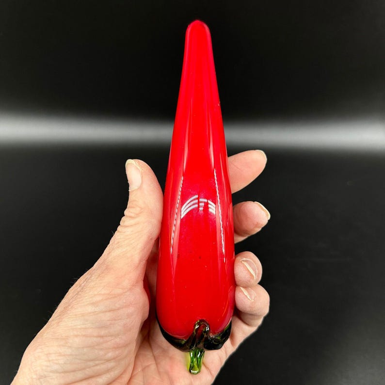 May include: A smooth, elongated red glass sculpture shaped like a chili pepper. The object features a glossy, vibrant red body that tapers to a point at the top, with a small, dark green glass stem detail at the base, reflecting light on its surface.