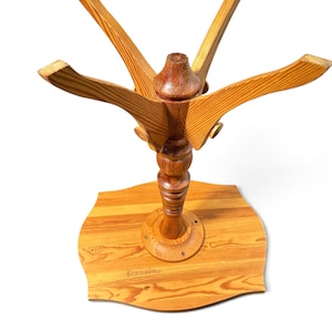 May include: A light brown wooden table base featuring a central turned pedestal column, four curved support arms extending outward, and a flat, four-sided base plate with rounded corners. The wood grain is visible throughout the structure.