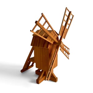 May include: A wooden model of a windmill featuring a vertical rectangular main structure with a sloped roof, four large lattice-style blades attached to a central hub, and a support base with angled legs extending outward from the bottom.