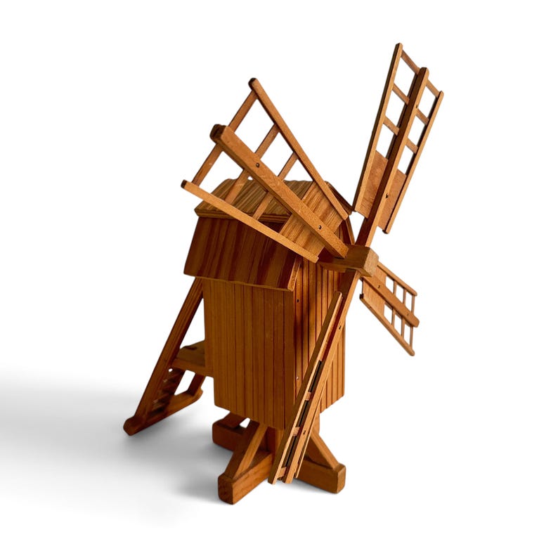 May include: A wooden model of a windmill featuring a vertical rectangular main structure with a sloped roof, four large lattice-style blades attached to a central hub, and a support base with angled legs extending outward from the bottom.