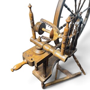 May include: A brown wooden spinning wheel featuring a large spoked wheel, a flyer assembly with a bobbin, and a horizontal frame supported by three turned legs. The structure includes a tensioning screw and a series of metal hooks on the flyer.
