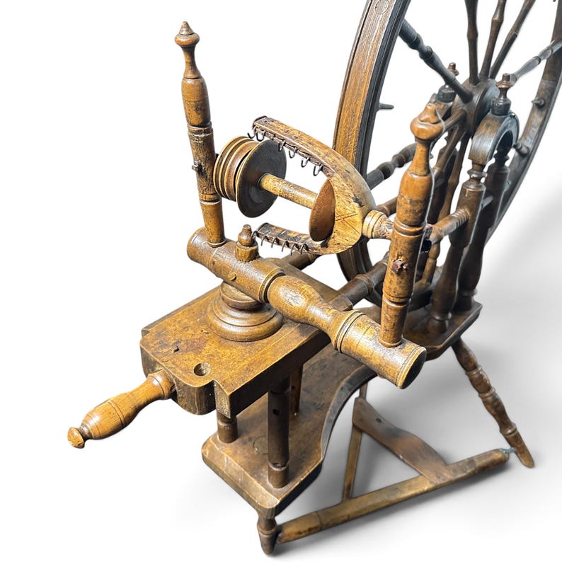 May include: A brown wooden spinning wheel featuring a large spoked wheel, a flyer assembly with a bobbin, and a horizontal frame supported by three turned legs. The structure includes a tensioning screw and a series of metal hooks on the flyer.