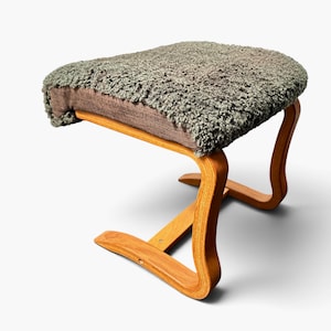 Puede incluir: Un reposapiés con una estructura de madera clara curvada y una base que se extiende hacia afuera. El asiento está tapizado con un material texturizado de piel sintética color verde salvia que envuelve los bordes, revelando una base de tela marrón.