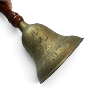 May include: A brass hand bell featuring an etched leaf pattern on the exterior surface. The bell is attached to a smooth, dark brown wooden handle with a rounded, turned design at the top of the metal bell section.