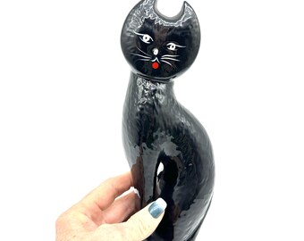 May include: A glossy black ceramic cat figurine with a tall, slender, curved body. The cat has painted white eyes, white whiskers, and a small red dot for a nose. The surface of the figurine has a textured, dimpled finish throughout the black glaze.