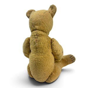 Pode incluir: Um urso de peluche de estilo vintage feito de tecido castanho texturizado, visto de costas. O urso apresenta costuras vis&iacute;veis ao longo do centro das costas e linhas horizontais escuras cosidas na sola do p&eacute; estendido.
