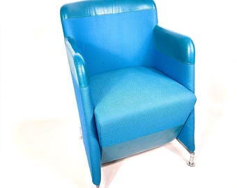 May include: An armchair featuring a bright blue fabric seat and backrest, accented with teal leather trim along the top edge and armrests. The chair is supported by four slender, polished silver metal legs with small circular feet at the base.