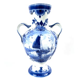 Op de afbeelding: Een wit en blauw keramische vaas met twee handvatten. De vaas heeft een gedetailleerde schildering van een zeilboot op een water. De vaas heeft een uitlopende rand en een voetstuk, met blauwe bloemaccenten.