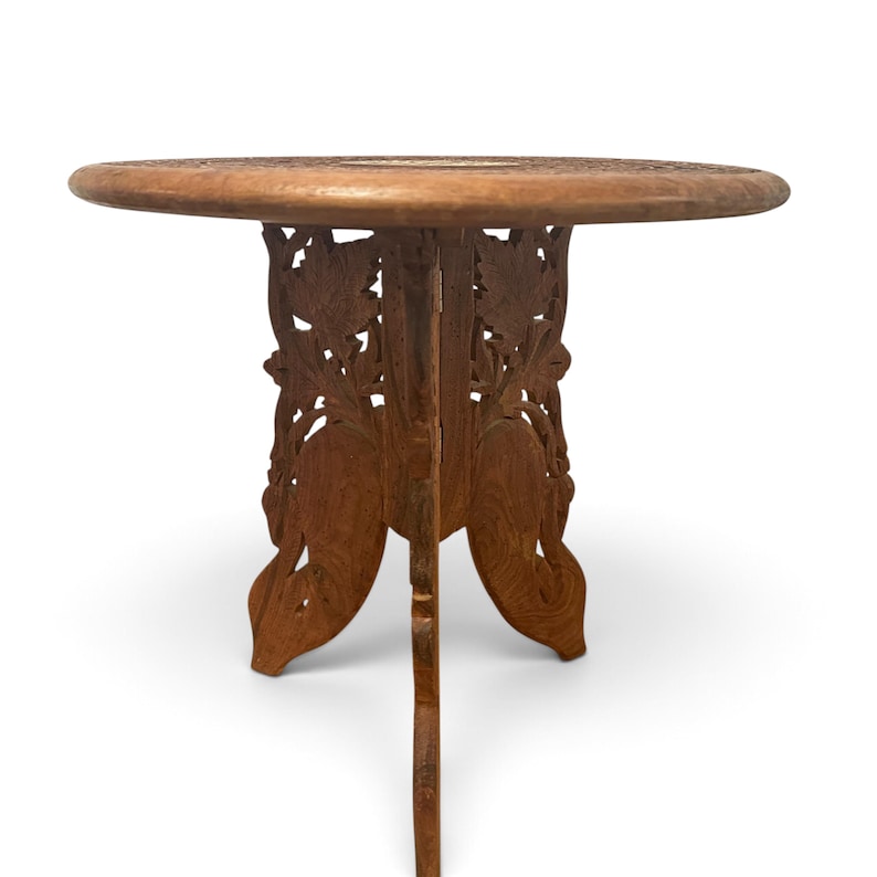 May include: A round wooden side table featuring a flat circular top and a folding base. The base is constructed from two interlocking panels intricately carved with leaf and floral patterns, providing a stable support for the table top.