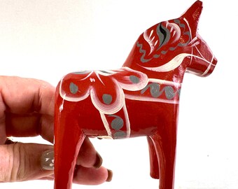 May include: A red wooden horse figurine featuring hand-painted white and grey floral patterns across its body, mane, and saddle area. The horse stands on four legs with a smooth, glossy finish and detailed decorative brushwork throughout its form.