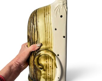 May include: A tall, flat ceramic cat figurine featuring a two-tone design. The left side has a textured, olive-green glaze with vertical lines, while the right side is smooth, off-white ceramic with small, dark, circular spots and etched whiskers.