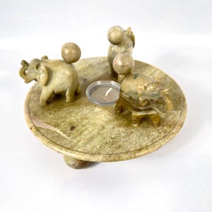 May include: A decorative candle holder featuring a round, stone base with three small elephant figurines and three spherical accents. A small glass candle holder is in the center. The candle holder is made of a light green and tan stone.
