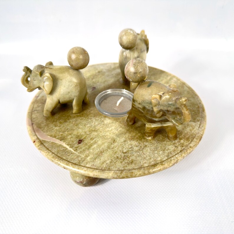 May include: A decorative candle holder featuring a round, stone base with three small elephant figurines and three spherical accents. A small glass candle holder is in the center. The candle holder is made of a light green and tan stone.