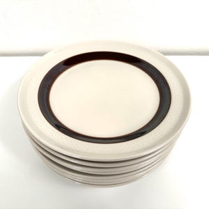 May include: A stack of six white ceramic dessert plates with a brown band and a white embossed pattern around the edge.