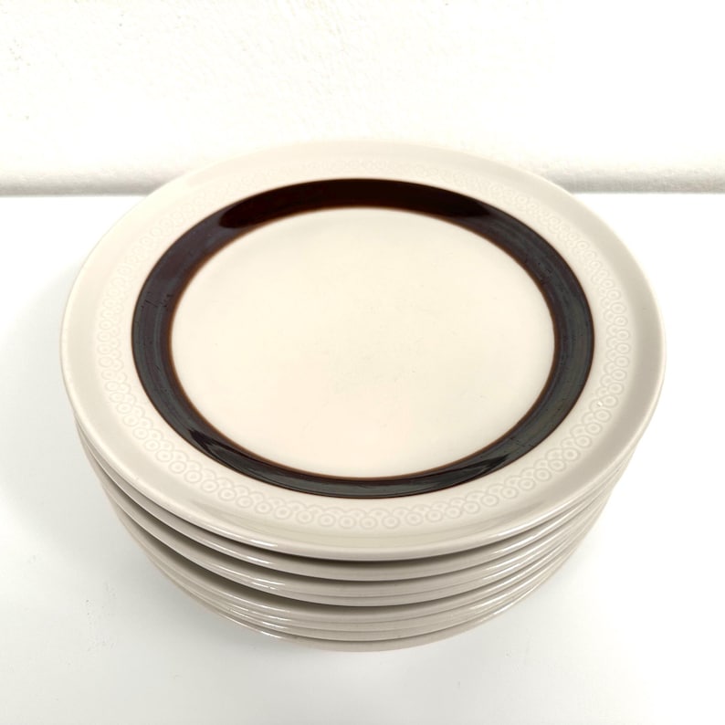 May include: A stack of six white ceramic dessert plates with a brown band and a white embossed pattern around the edge.