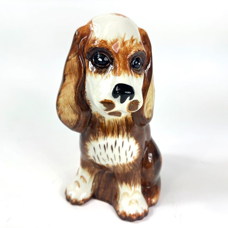 Peut inclure: Figurine en c&eacute;ramique repr&eacute;sentant un chien, avec un sch&eacute;ma de couleurs marron et blanc. Le chien a de grands yeux expressifs et des oreilles tombantes. La figurine est en position assise, avec ses pattes visibles. Le pelage du chien est d&eacute;taill&eacute; avec des marques marron et blanches.