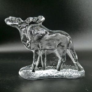 May include: A clear glass sculpture of a moose standing on a textured base. The moose has detailed antlers and a realistic form. The sculpture is transparent, allowing light to pass through.