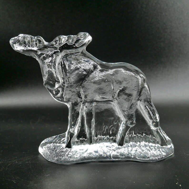May include: A clear glass sculpture of a moose standing on a textured base. The moose has detailed antlers and a realistic form. The sculpture is transparent, allowing light to pass through.