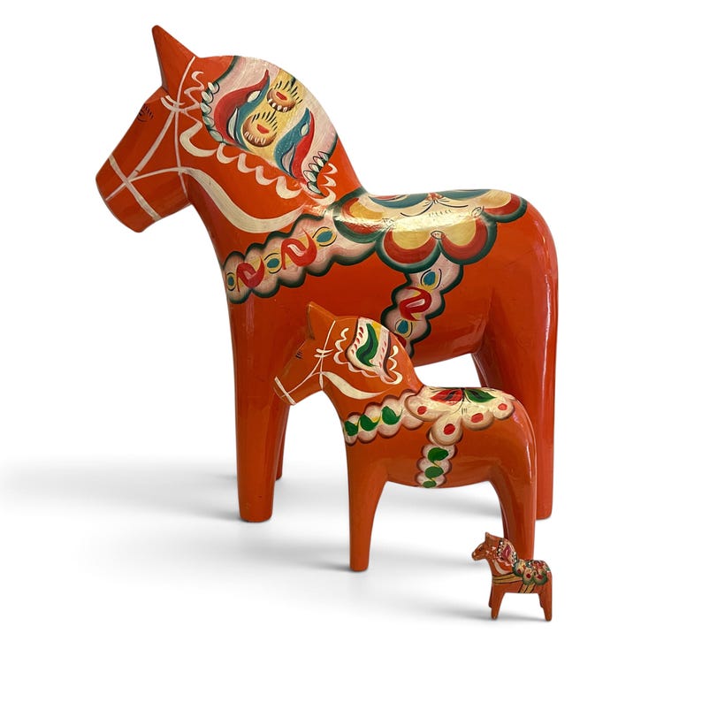 Puede incluir: Tres figuras de caballos Dala de color naranja en diferentes tama&ntilde;os, cada una con patrones florales y geom&eacute;tricos pintados a mano en blanco, verde, azul y dorado. Los caballos est&aacute;n dispuestos en orden descendente de tama&ntilde;o con un acabado brillante.