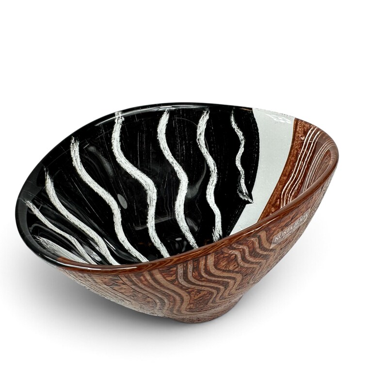 May include: A glass bowl featuring a black interior with white vertical wavy lines and a white section. The exterior has a brown textured pattern with wavy lines. A small rectangular label with the text Kosta Boda is visible on the side.