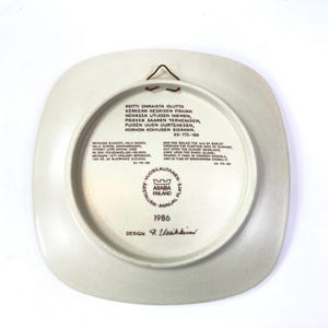 Op de afbeelding: Een vierkante, gebroken witte keramische plaat met een bruine metalen hanger aan de achterkant. De plaat bevat tekst in het Fins en Engels, samen met het jaar 1986 en het Arabia Finland-logo. Het ontwerp van de plaat bevat een gedicht.
