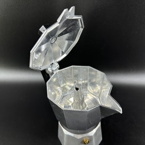 Puede incluir: Una cafetera de espresso de aluminio plateado con la tapa abierta, revelando los componentes internos. La cafetera tiene una base redonda y un pico en el lateral.