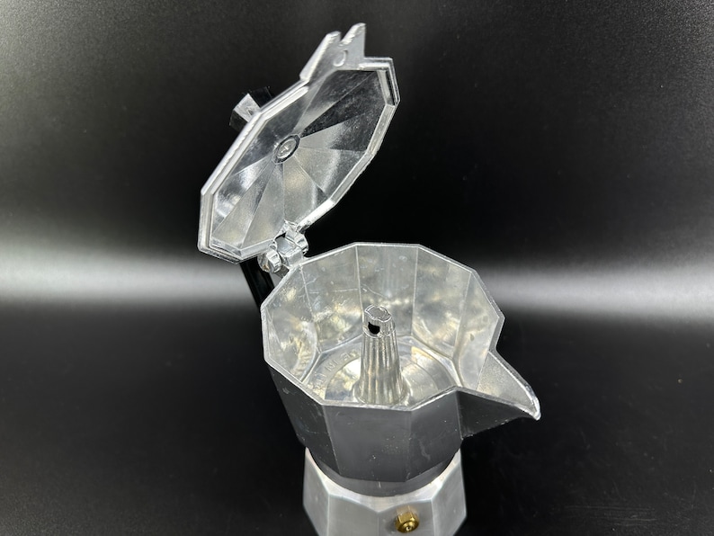 Puede incluir: Una cafetera de espresso de aluminio plateado con la tapa abierta, revelando los componentes internos. La cafetera tiene una base redonda y un pico en el lateral.