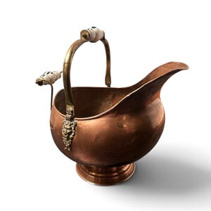 Pu&ograve; includere: Un secchiello per carbone in rame con finitura lucida, dotato di un manico in ottone con inserti in ceramica bianca. Il contenitore presenta un ampio beccuccio, una base circolare a piedistallo e decorazioni in ottone a testa di leone.