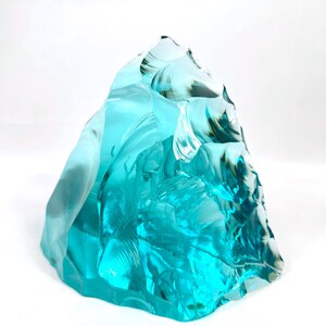 May include: A large, irregularly shaped sculpture made of translucent, aqua-colored glass. The glass has a rough, textured surface with areas of darker green and blue. The sculpture is set against a white background.