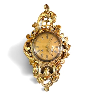 May include: A gold-colored wall clock featuring an ornate, carved frame with intricate scrollwork, floral motifs, and a lattice pattern. The clock face displays black Roman numerals, Arabic minute markers, and decorative metal hands behind glass.