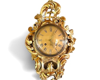 Puede incluir: Un reloj de pared de color dorado con un marco tallado y ornamentado que presenta intrincados motivos de volutas, flores y un patrón de celosía. La esfera muestra números romanos negros, marcas de minutos arábigas y manecillas de metal.