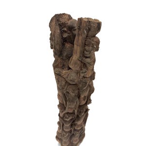 May include: A vertical dark brown wooden sculpture featuring a dense, interconnected carving of multiple human figures stacked and intertwined from top to bottom. The textured surface displays intricate details of faces and limbs carved into the wood.