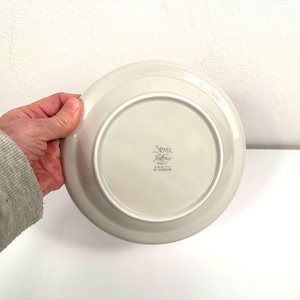 May include: White ceramic dinner plate with a raised rim. The bottom of the plate has the words "Orma" and "Made in Italy" printed in black.