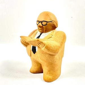 Könnte beinhalten: Beigefarbene Figur eines Mannes im Anzug, der ein Buch liest. Die Figur hat eine schwarze Brille und eine schwarze Krawatte. Die Figur steht auf einer weißen Oberfläche.