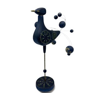 May include: A dark blue wooden bird figurine standing on a thin metal rod and a circular base. The bird features painted green and white circular patterns on its body, a gold-colored beak, and a tail made of thin wires tipped with blue spheres.