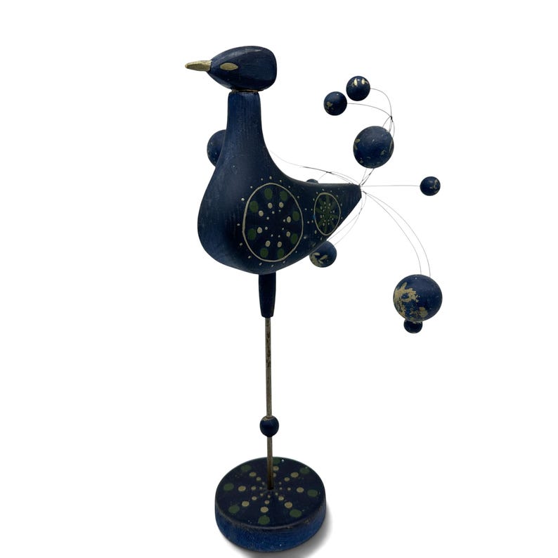 May include: A dark blue wooden bird figurine standing on a thin metal rod and a circular base. The bird features painted green and white circular patterns on its body, a gold-colored beak, and a tail made of thin wires tipped with blue spheres.