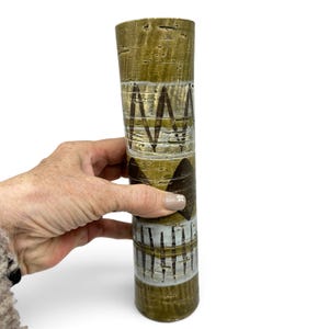 Op de afbeelding: Een hoge cilindrische keramische vaas met een textuurrijke olijfgroene glazuurlaag en horizontale banden van witte en donkerbruine geometrische patronen, waaronder zigzaglijnen en massieve driehoekige vormen in een verticaal ontwerp.