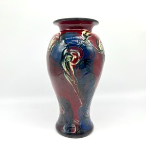May include: A tall, decorative vase with a flared base and a narrow neck. The vase features a glossy finish with a blend of red, blue, and cream-colored swirling patterns. The rim and base are dark, creating a striking contrast.