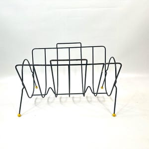 May include: Black metal magazine rack with a mid-century modern design. The rack features a series of vertical and curved wire supports, with yellow ball feet. The rack is designed to hold magazines and other reading materials.