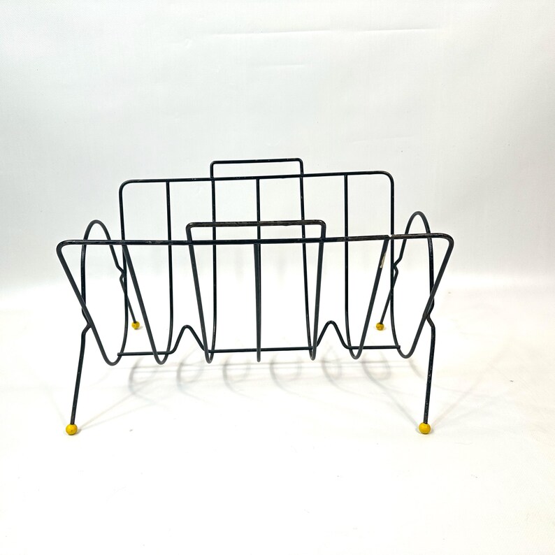 May include: Black metal magazine rack with a mid-century modern design. The rack features a series of vertical and curved wire supports, with yellow ball feet. The rack is designed to hold magazines and other reading materials.