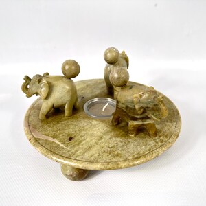 May include: A round, light green stone candle holder with four carved elephants and a small candle. The elephants are positioned around the edge of the holder, and a small candle is in the center. The candle holder is approximately 5 inches in diameter.