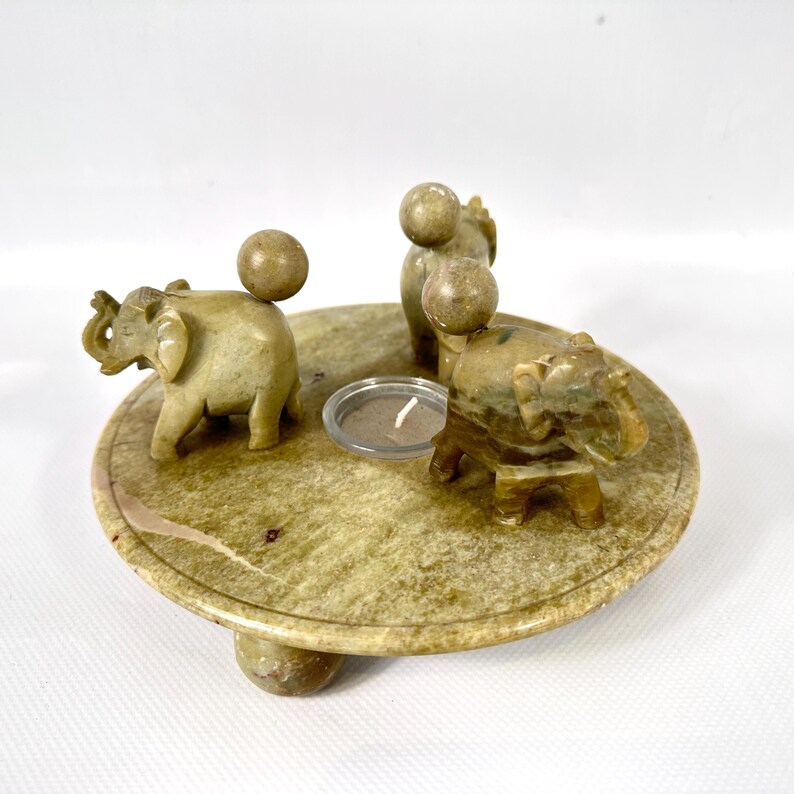 May include: A round, light green stone candle holder with four carved elephants and a small candle. The elephants are positioned around the edge of the holder, and a small candle is in the center. The candle holder is approximately 5 inches in diameter.