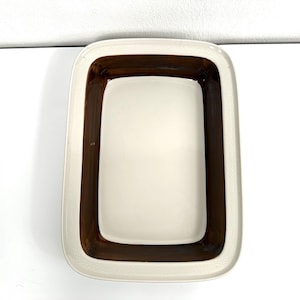 Puede incluir: Una fuente rectangular de cerámica para horno con una base central lisa de color crema rodeada por un borde ancho esmaltado en marrón oscuro. El borde exterior presenta un sutil patrón geométrico en relieve de color crema.