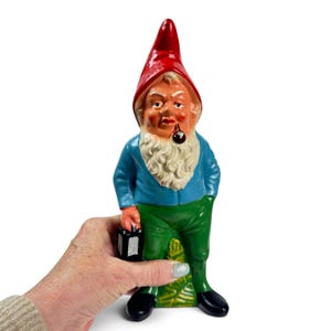 May include: A garden gnome figurine featuring a tall red pointed hat, a blue long-sleeved shirt, green trousers, and black shoes. The gnome has a white beard, a painted face, and holds a small black lantern in one hand while a pipe is in its mouth.