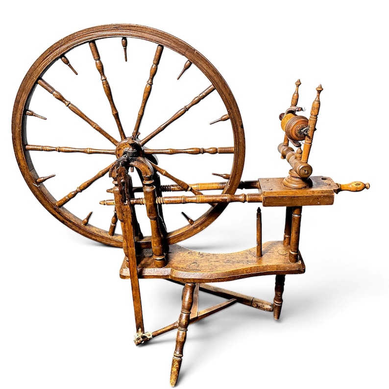 May include: A brown wooden spinning wheel featuring a large circular wheel with numerous spokes, a horizontal bench, a vertical flyer assembly with a bobbin, and a tripod base. The structure is crafted from turned wood components throughout.