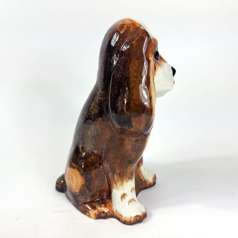Peut inclure: Figurine en c&eacute;ramique repr&eacute;sentant un chien assis, avec une finition brillante. Le chien est brun et blanc, avec de longues oreilles tombantes et un visage d&eacute;taill&eacute;. Objet d&eacute;coratif.